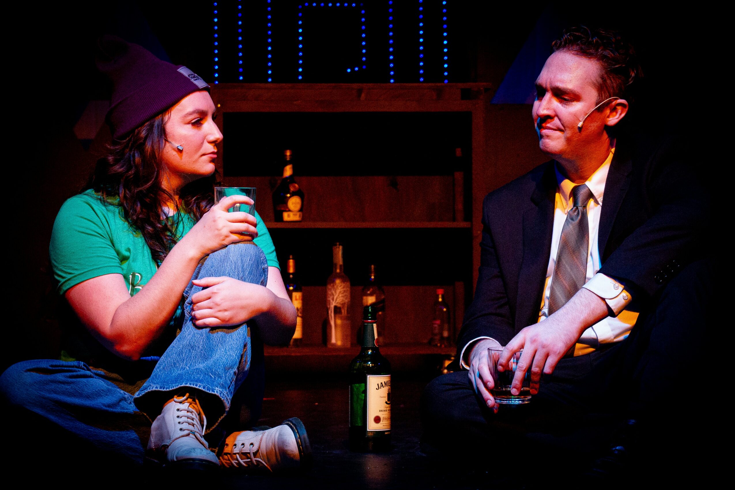 Two people sitting and talking in a dimly lit room with bottles on a shelf behind them.