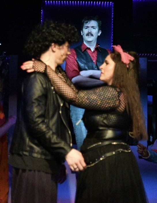 Two people embrace in a dark setting with a man in a framed portrait in the background.