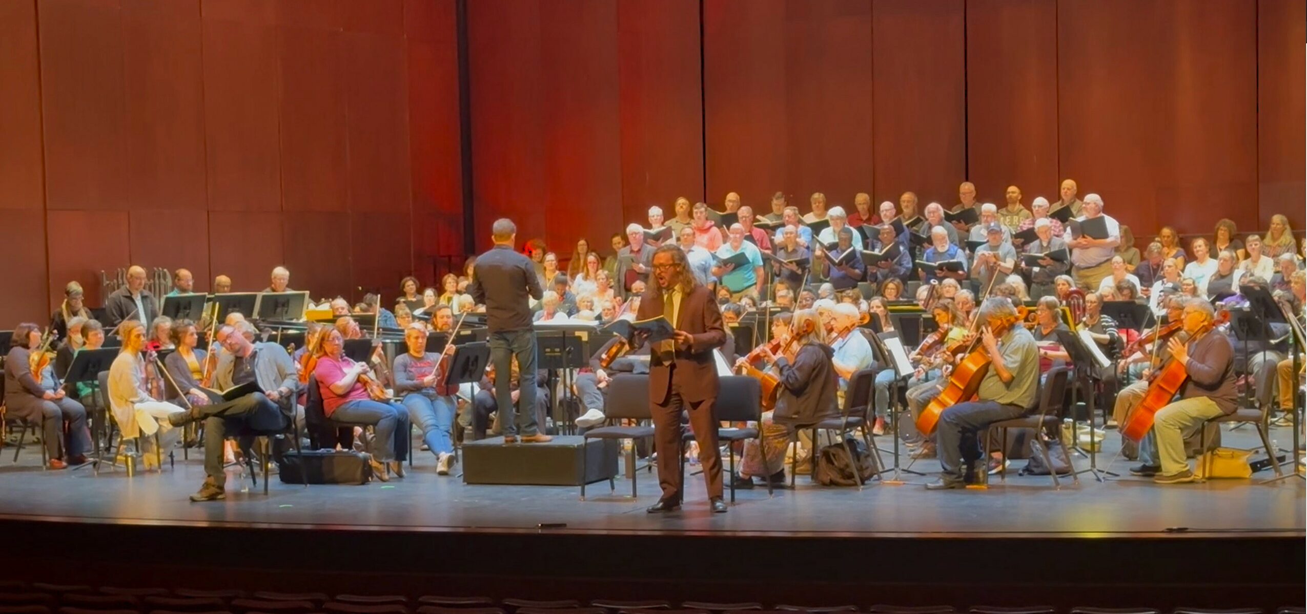 Large choir or orchestra performing on stage with a conductor, audience seated in the background, wooden wall backdrop.