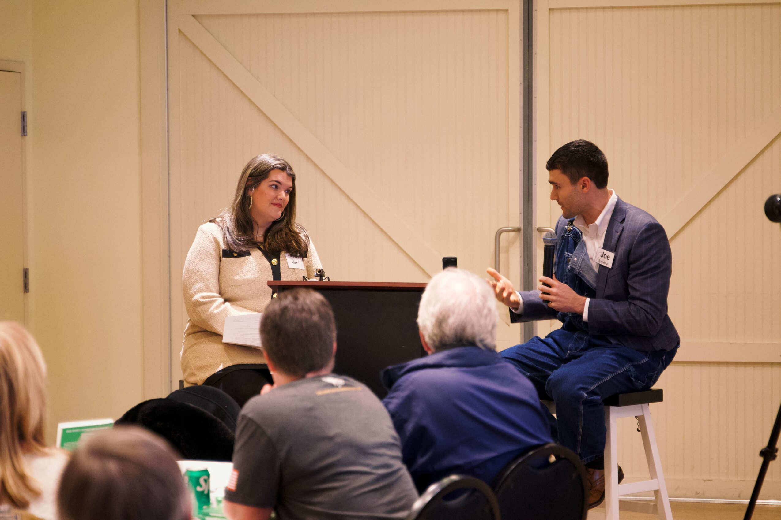Two people sit on stools on stage, engaging in a conversation, with an audience watching. The background is a plain wall.