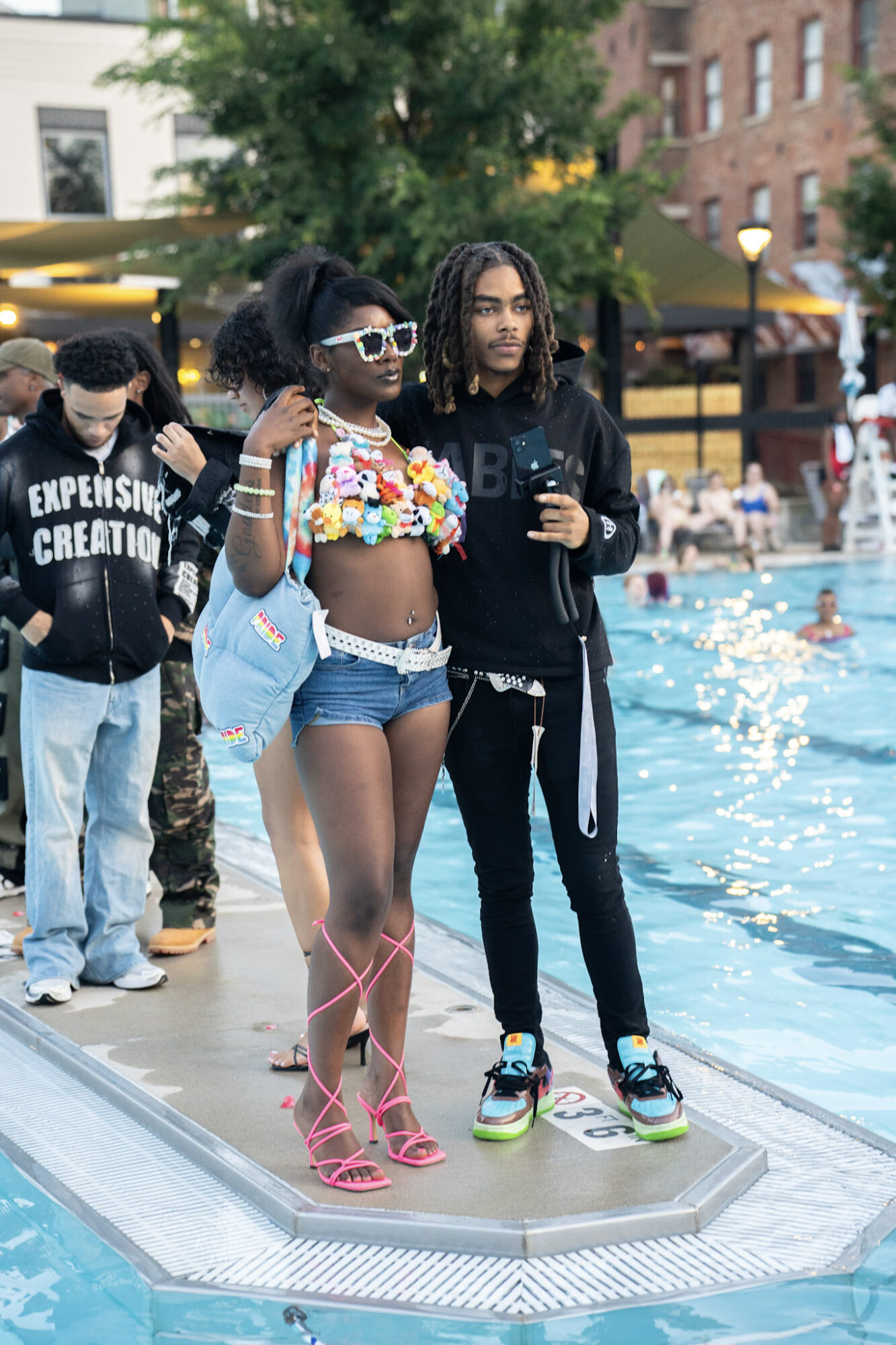 Two young women stand by a poolside, one in colorful crop top and shorts, the other in dark hoodie and pants, with people in the background.