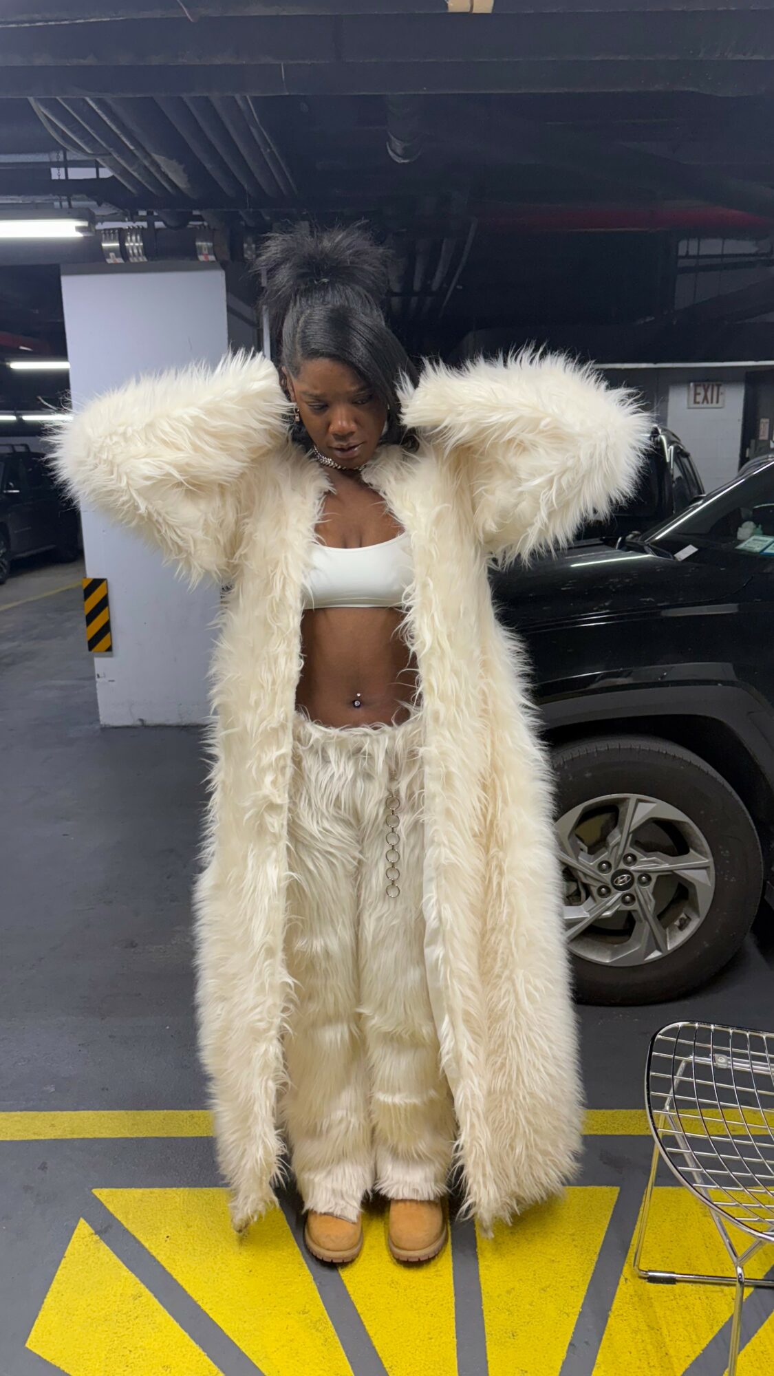 Person standing in parking garage wearing a long fluffy coat, crop top, and pants, with a black vehicle behind.