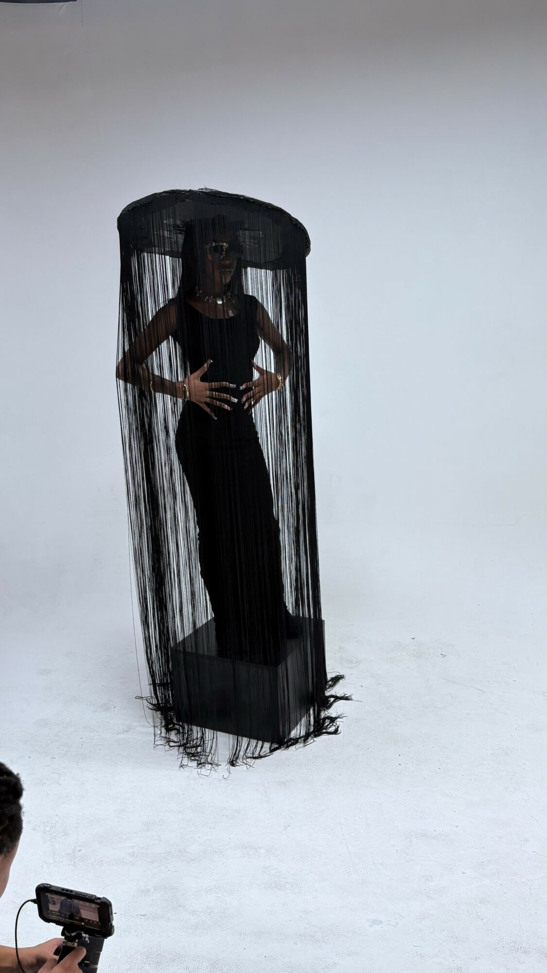 Person standing inside a tall, black, cage-like structure with a sheer curtain, on a plain white background.
