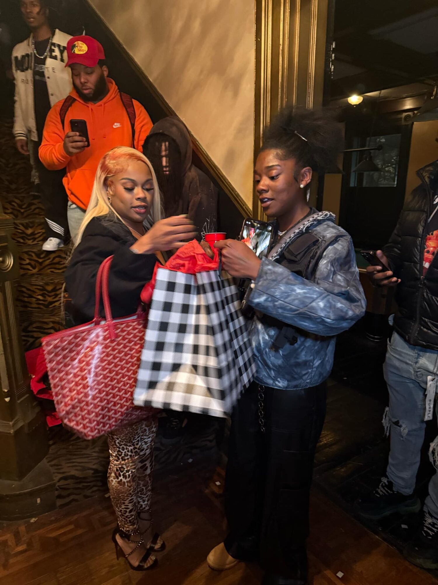 Two women stand indoors, one with blonde hair and leopard print pants, holding shopping bags, and the other with dark hair in a ponytail, wearing a denim jacket, holding a gift box.