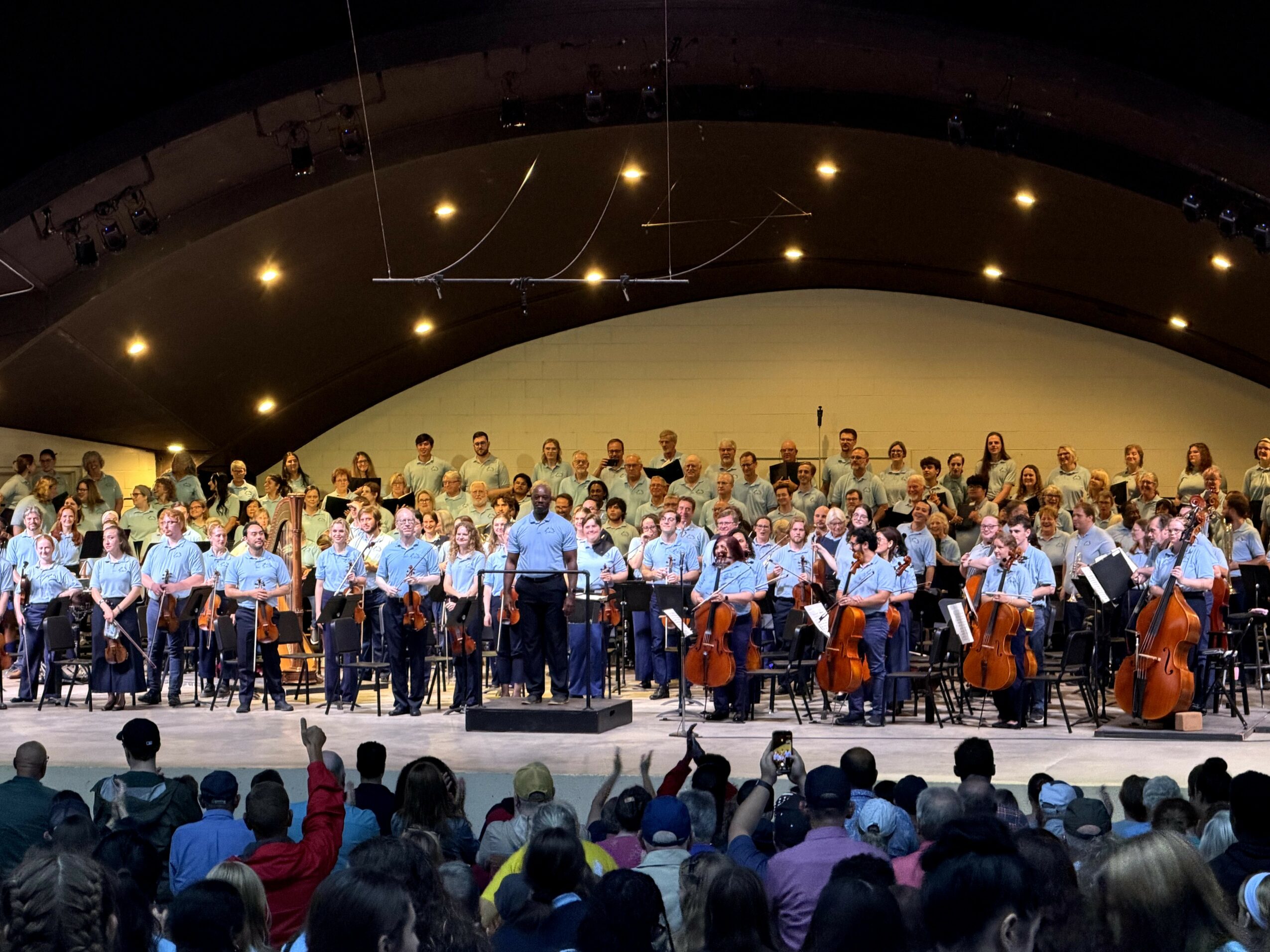 Orchestra and choir perform on stage with audience watching, under a curved ceiling with lights.