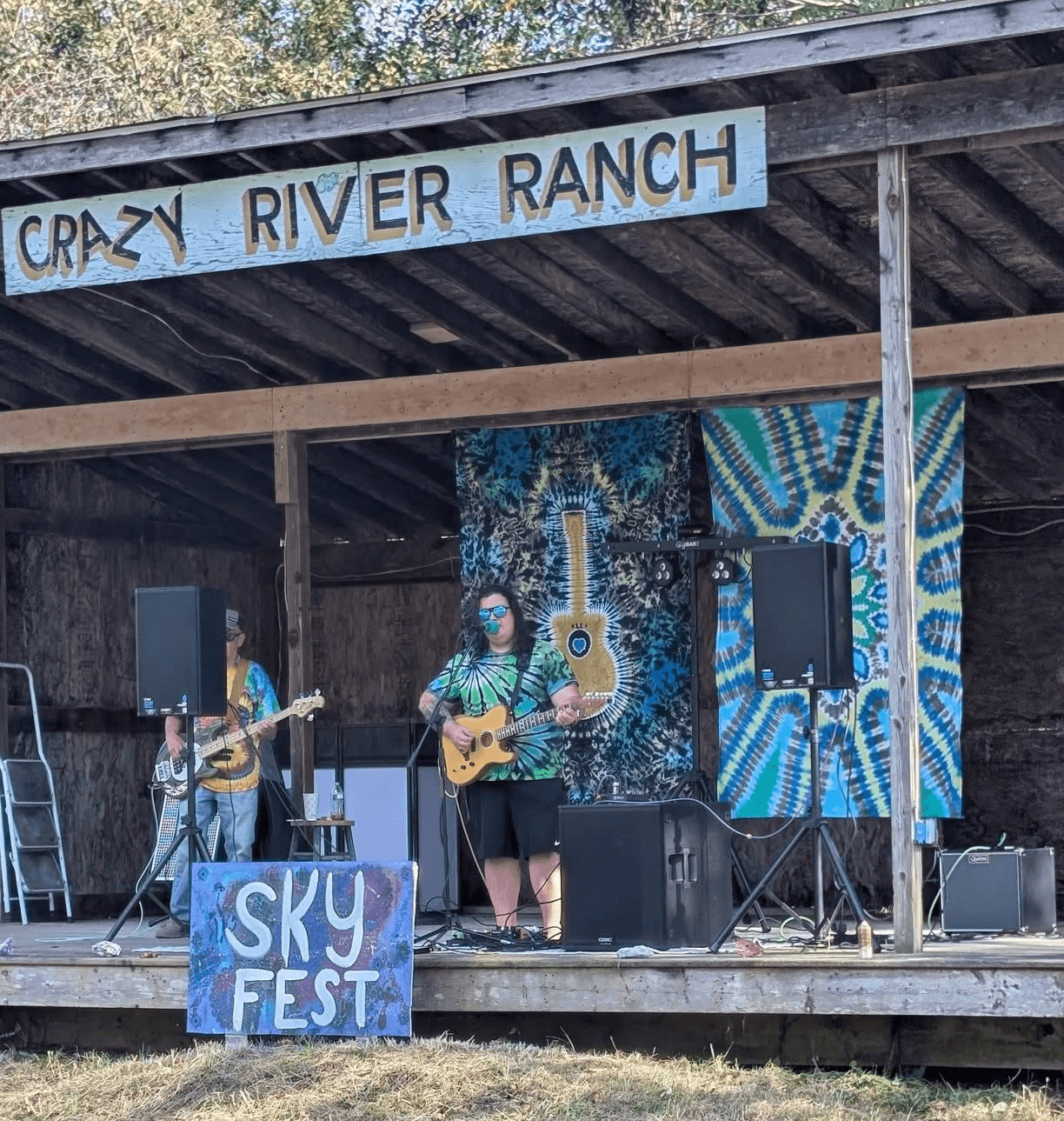 Three people perform on an outdoor stage with a sign reading 'Crazy River Ranch' and a 'Sky Fest' sign, playing guitars.