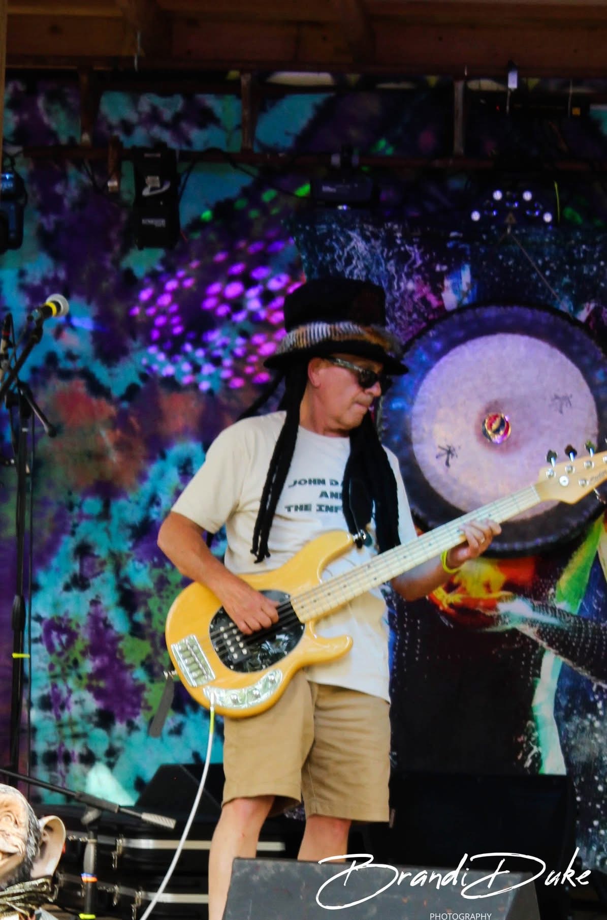 Person playing electric guitar on stage with colorful cosmic background, wearing sunglasses, hat, and casual clothing.