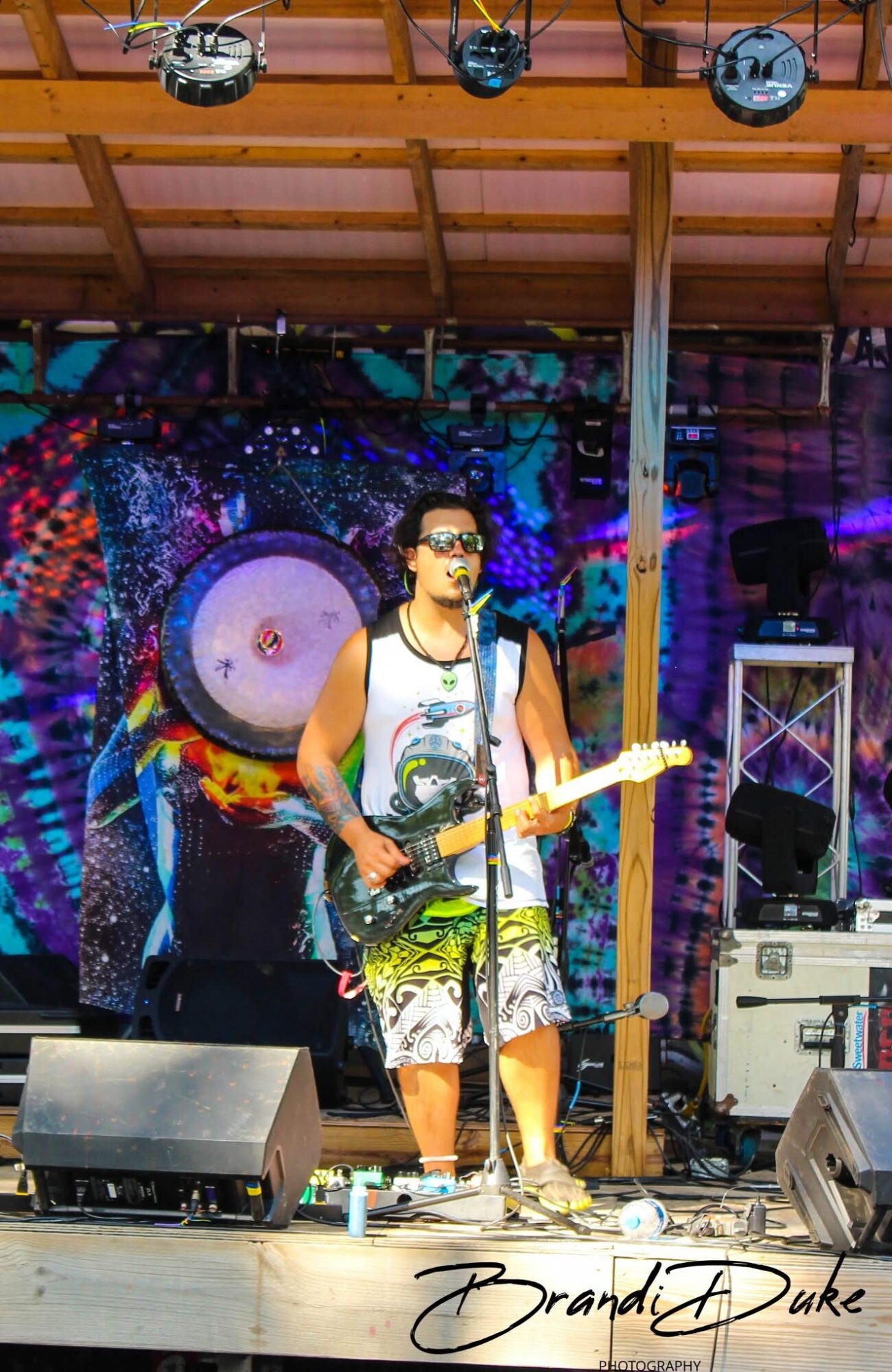 A man wearing sunglasses, a tank top, and shorts playing an electric guitar on stage with a colorful cosmic background.