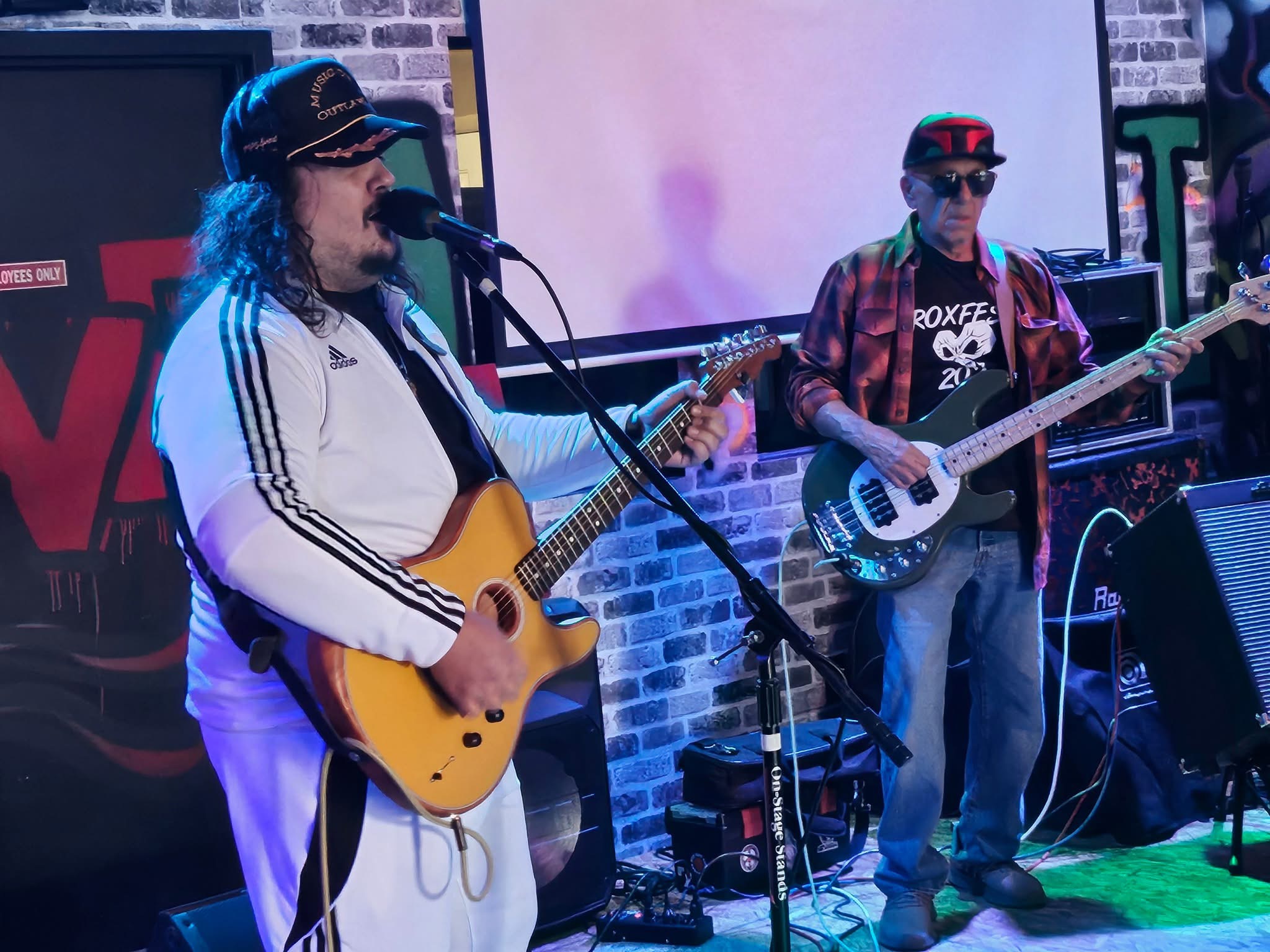 Two musicians perform on stage, one singing and playing guitar, the other playing bass guitar, with a brick wall background.