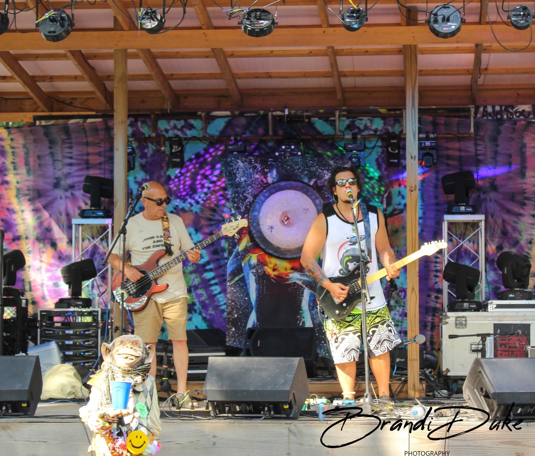 Two musicians perform on an outdoor stage with colorful background, one singing and playing guitar, the other playing bass.
