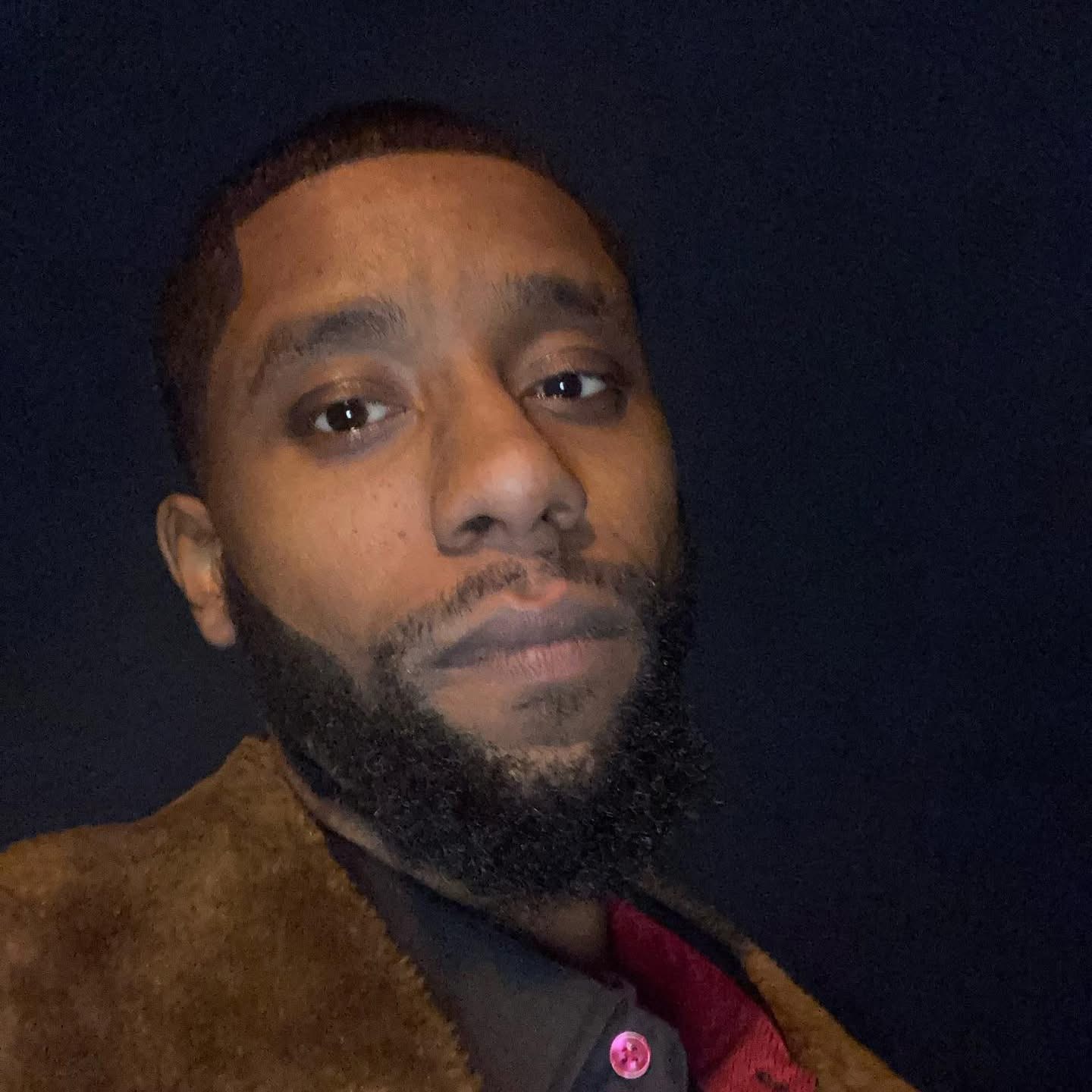 Close-up of a man with a beard and short hair, wearing a brown jacket and a dark shirt, against a dark background.