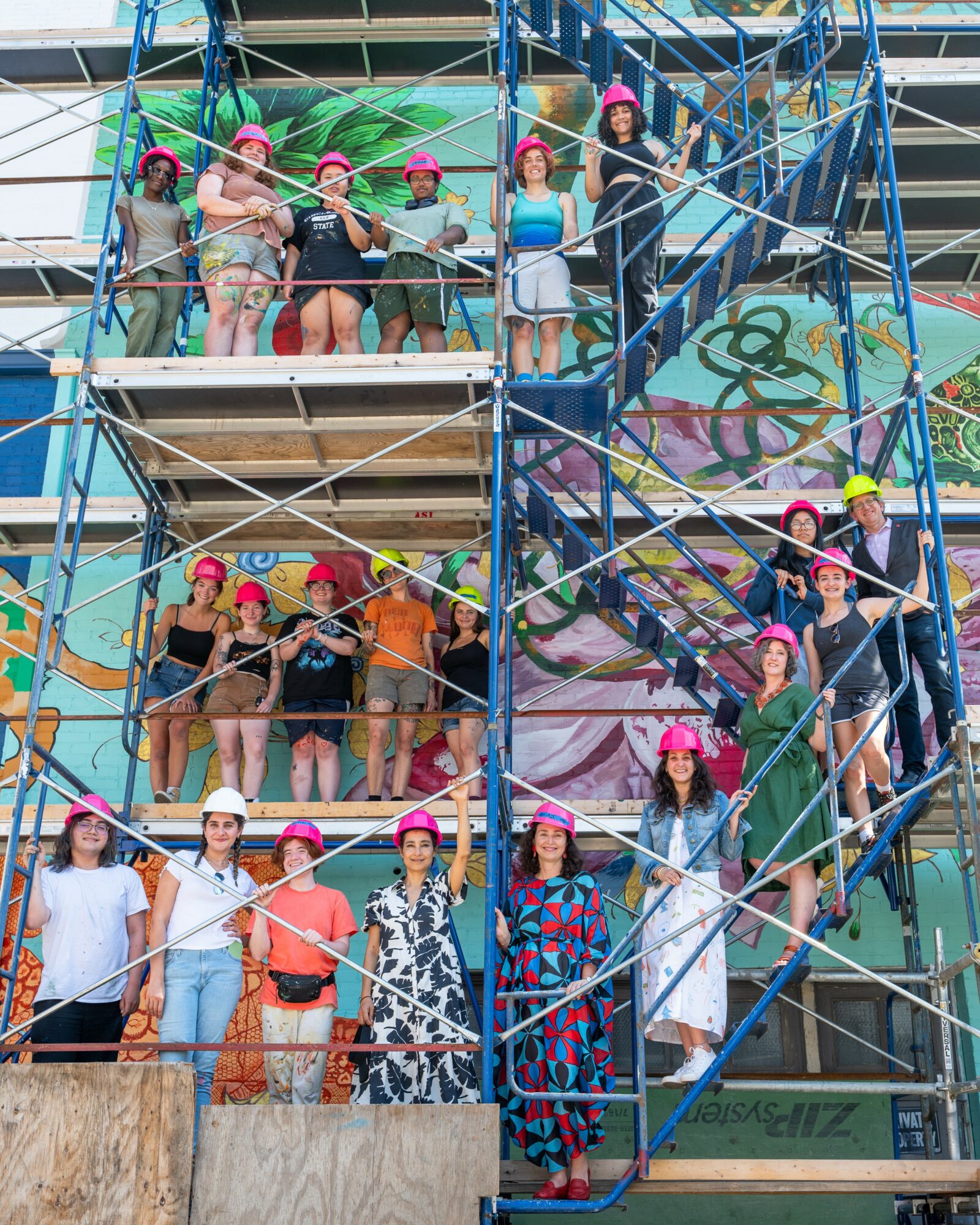 Group of people wearing pink hats standing on scaffolding in front of a colorful graffiti wall.