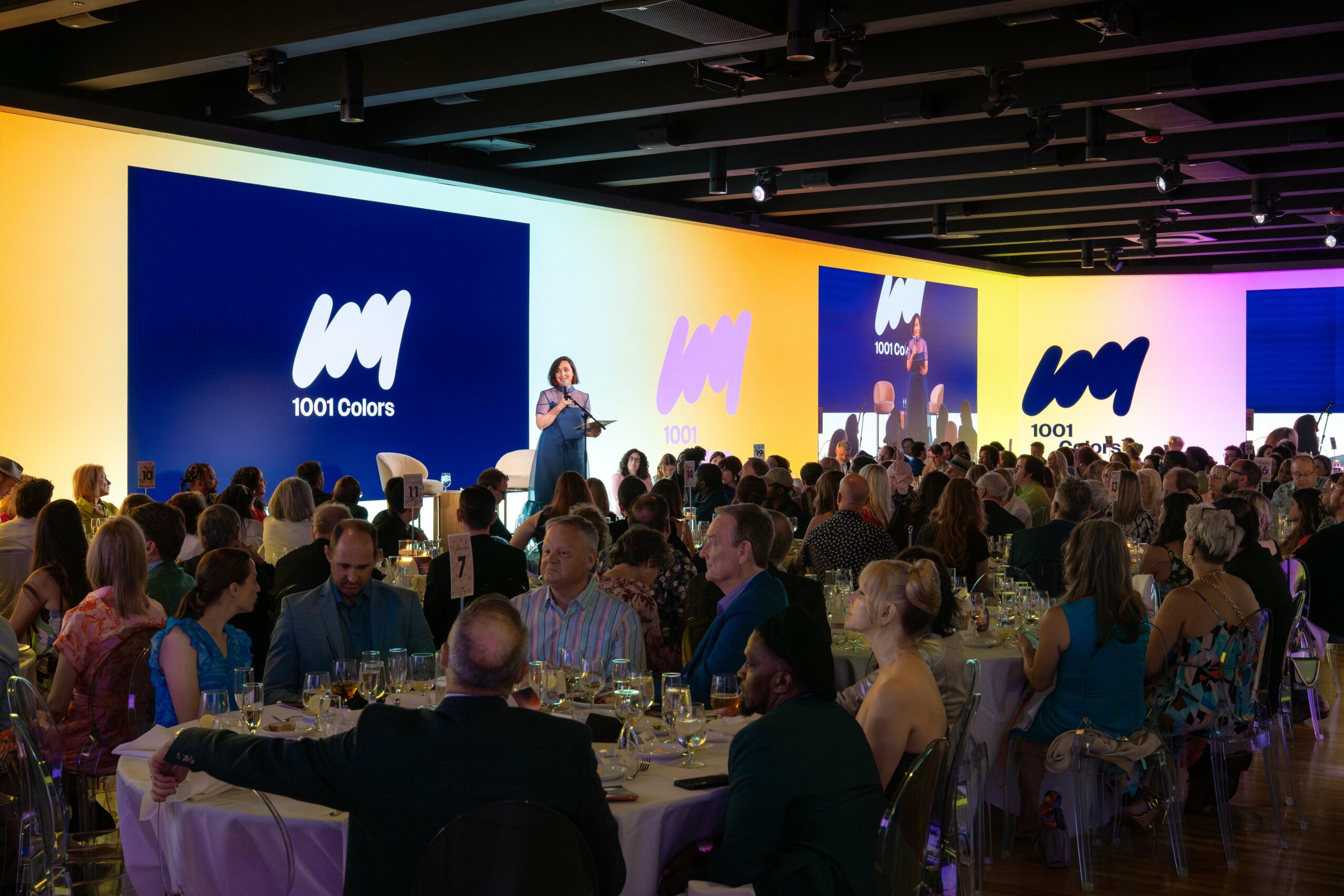 Large event with a stage, speaker, and audience seated at tables in a spacious hall.