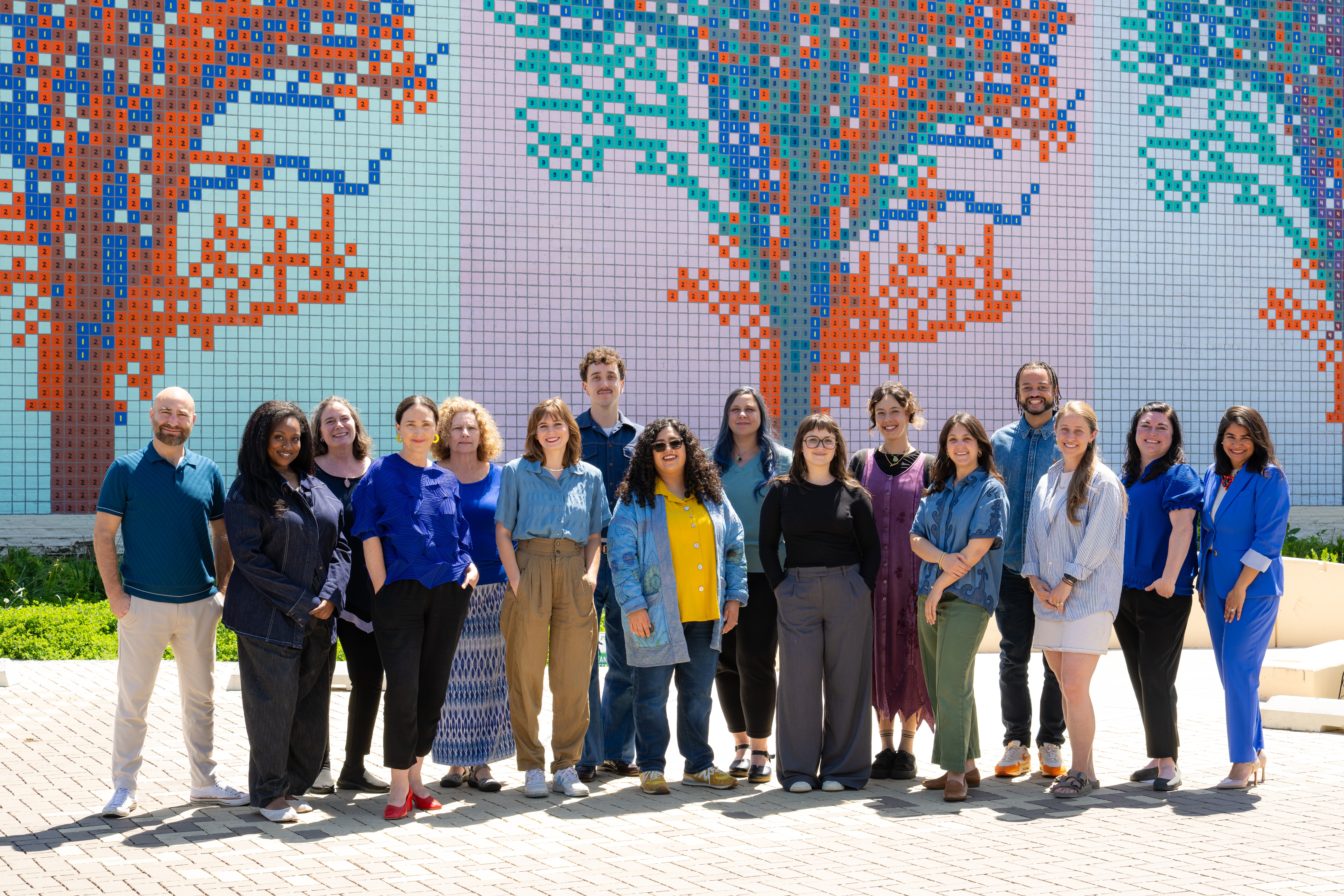 Group of 14 people standing outdoors in front of a colorful pixelated tree mural, smiling and posing for the photo.