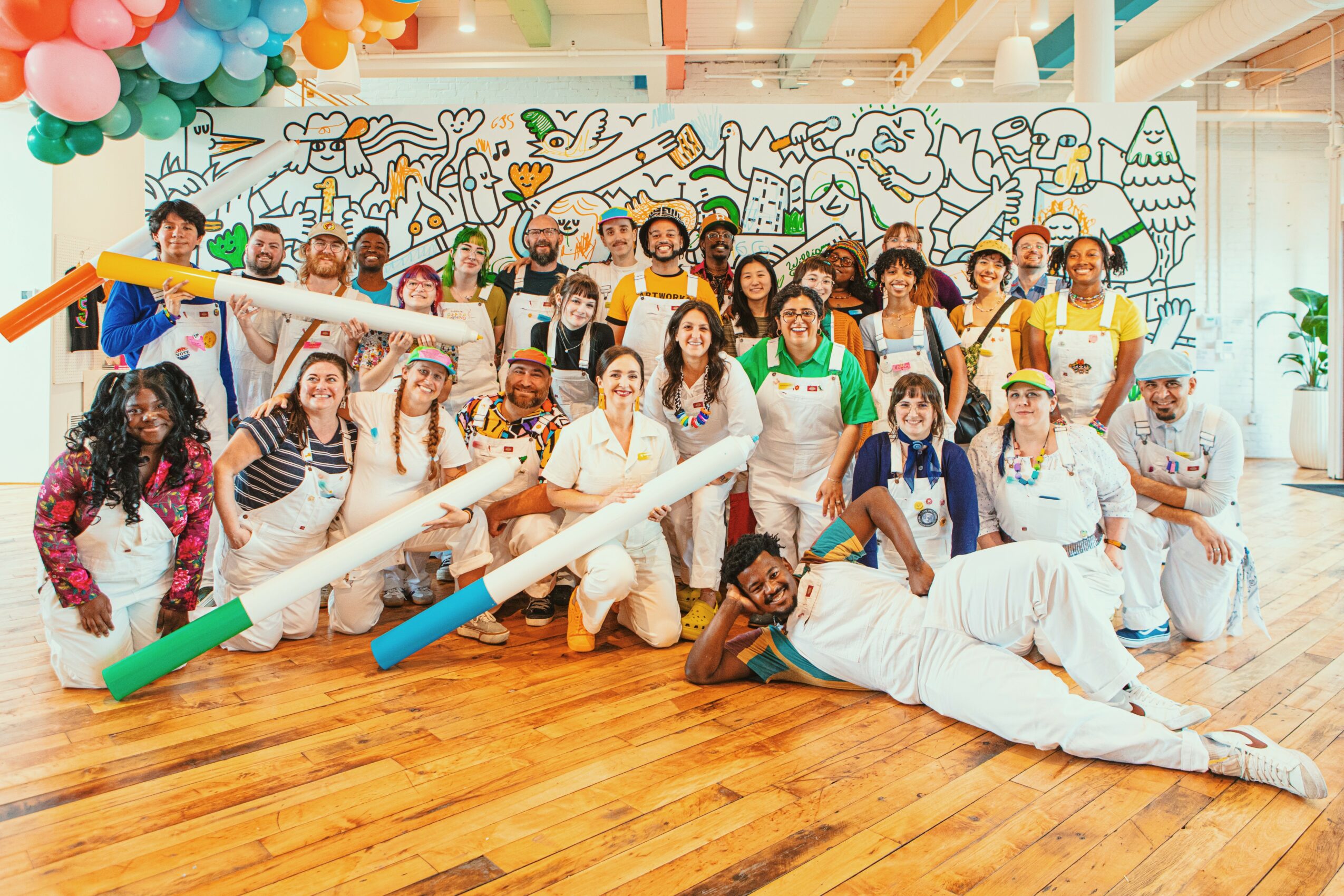 Group of people in colorful costumes and props, smiling in a decorated room with a mural background.