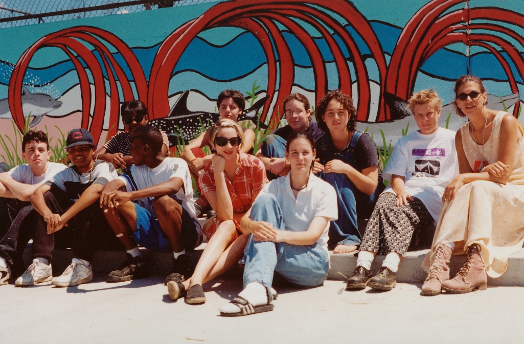 Group of ten people sitting outdoors in front of colorful red and blue abstract sculpture, some wearing sunglasses.