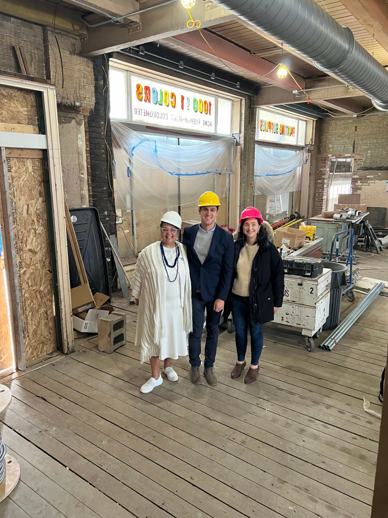 Three people standing inside a bakery or cafe, with a colorful sign and wooden flooring, in a casual setting.