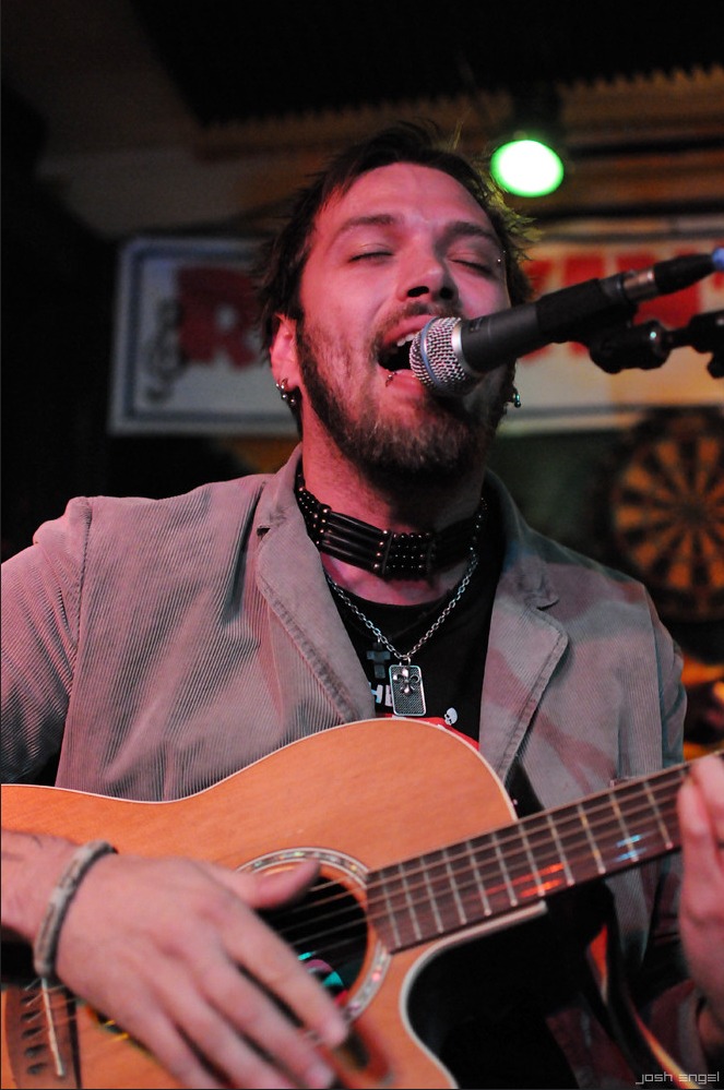 Male musician singing into a microphone while playing an acoustic guitar in a dimly lit venue.