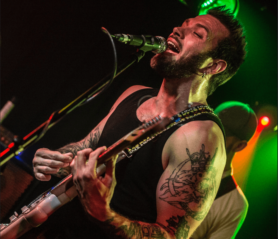 Male musician singing into microphone and playing guitar, tattoos visible on arm, stage lighting in background