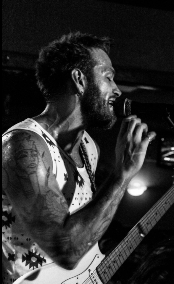 Man singing into a microphone while playing an electric guitar, with tattoos on his arm, in black and white.