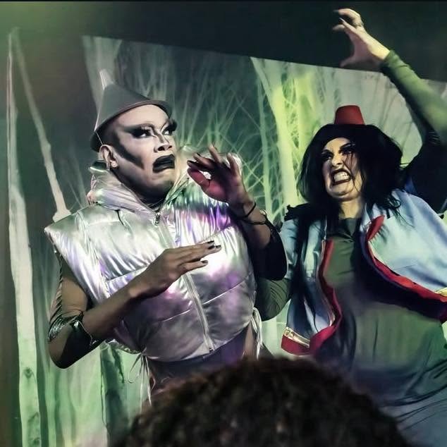 Two performers in colorful costumes and makeup on stage with green lighting background, one with a hat and the other with a cap, gesturing animatedly.