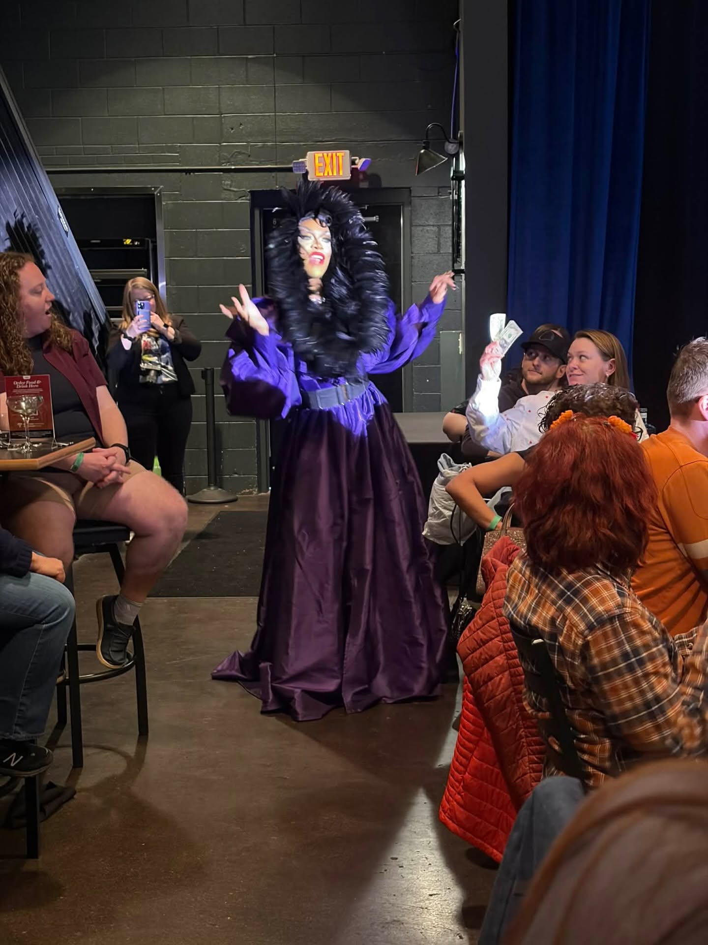 Performer dressed as a witch with a large black wig and purple gown stands in front of seated audience in a theater.