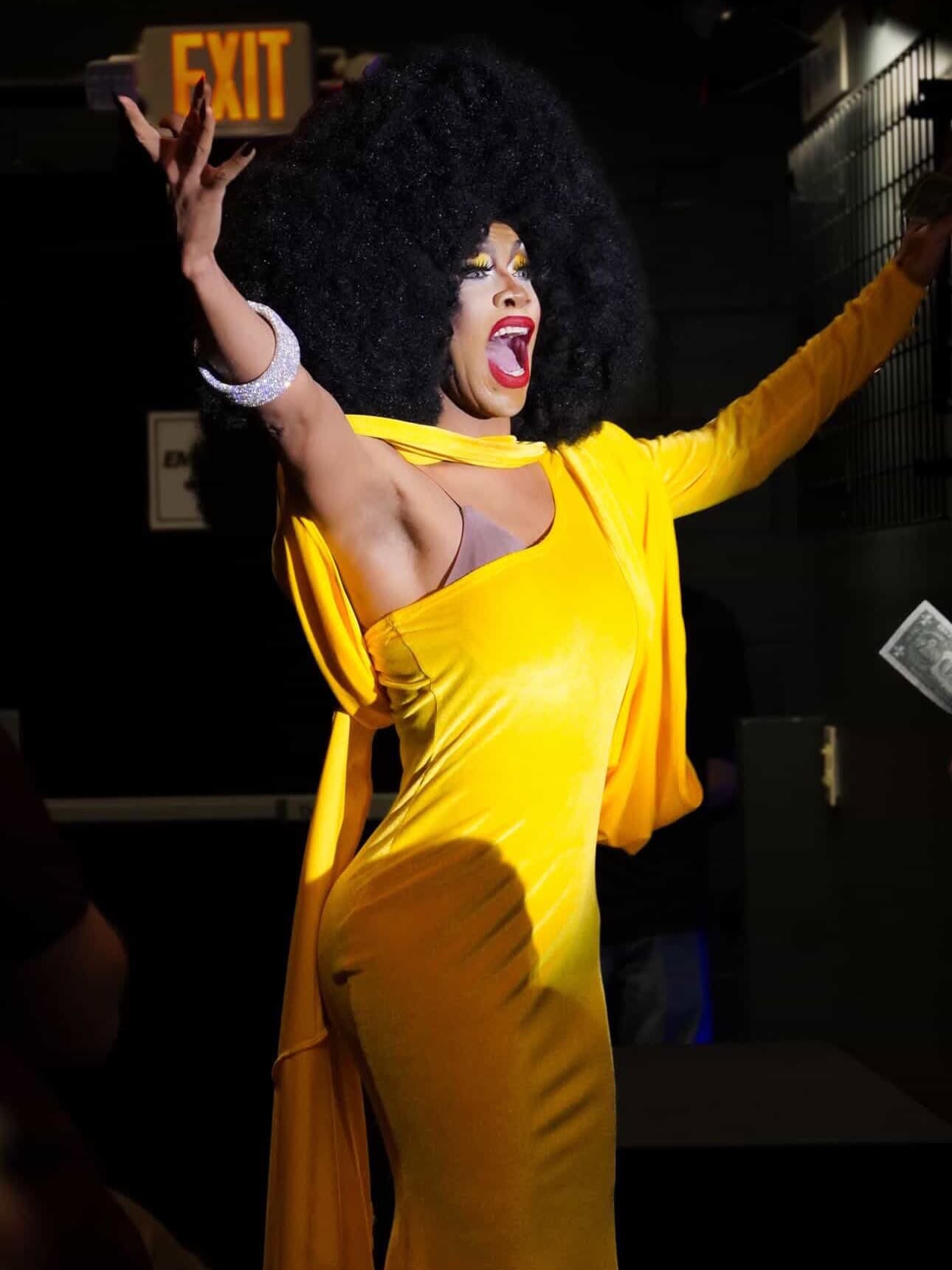 Woman with large curly hair in yellow outfit, arms raised, smiling, in a dark setting with an exit sign behind her.