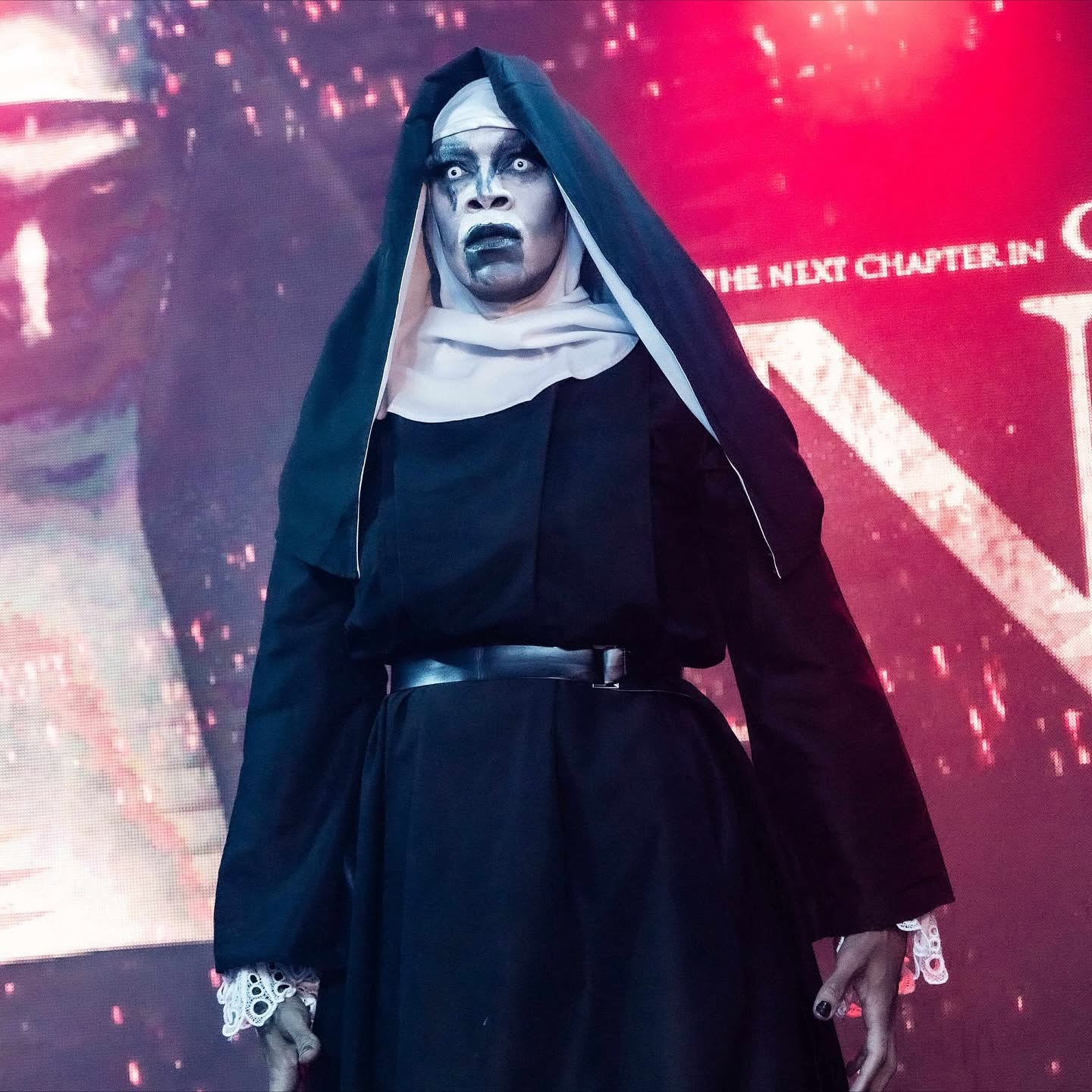 Person dressed as a nun with dark makeup and a black habit, standing against a red background.