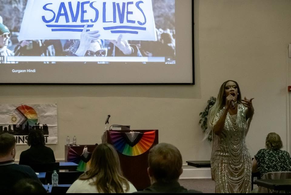 A woman speaks to an audience in a room with a large screen displaying a sign that says 'SAVES LIVES'.