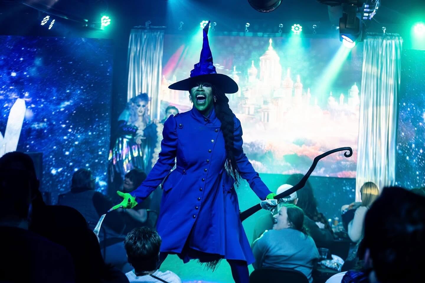 Person dressed as a witch with a hat and wand on stage during a performance, audience watching, colorful background.