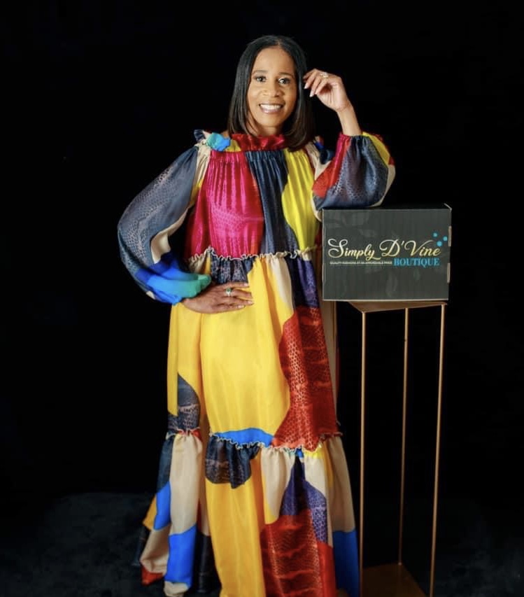 Smiling woman in colorful dress standing next to a sign that reads 'Simply D'Voie Boutique' against a black background.