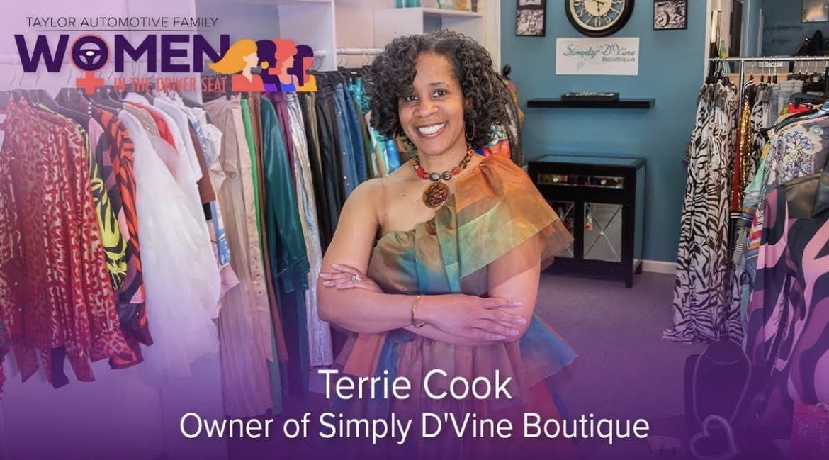 Woman with curly hair in colorful dress inside boutique, smiling, with clothing racks in background.