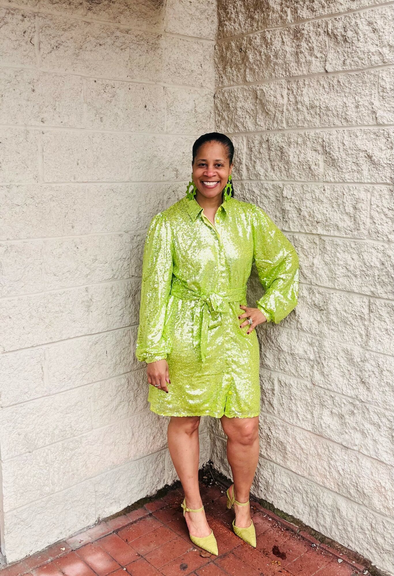 Woman in a shiny green dress and yellow heels standing against a textured beige wall, smiling, with one hand on her hip.