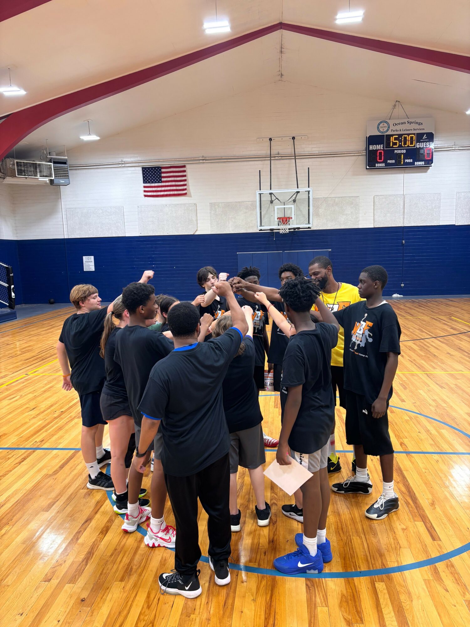 Group of people in a gymnasium, standing in a circle, engaging in a team activity or discussion.