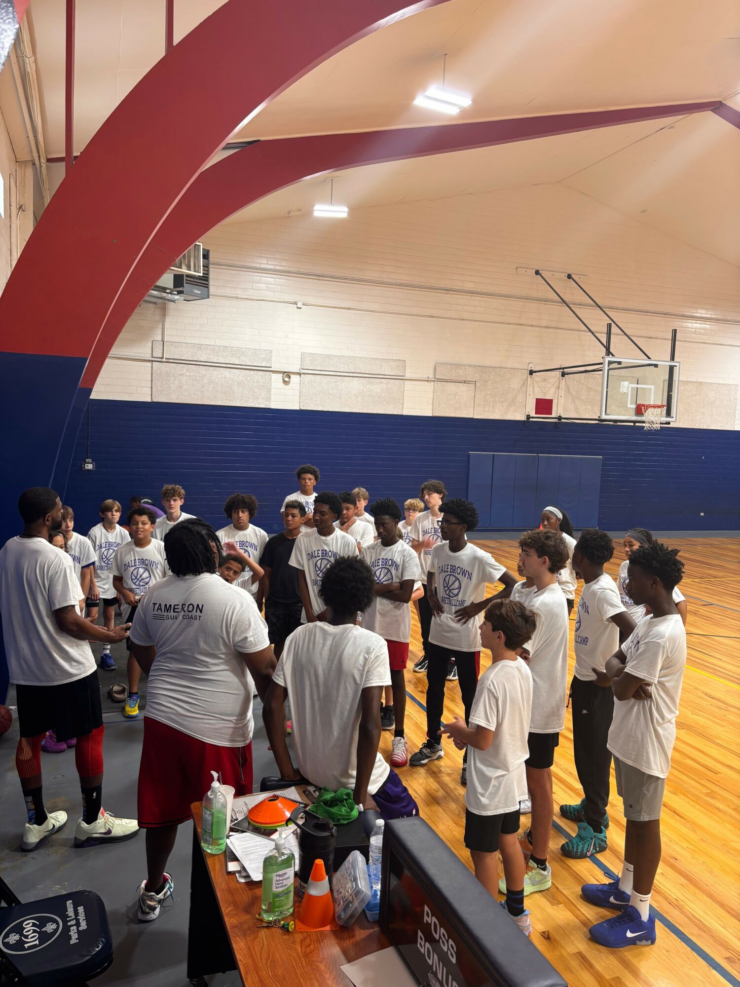 Group of children in white shirts gathered in a gymnasium near a basketball hoop, some sitting and some standing, with a table nearby.