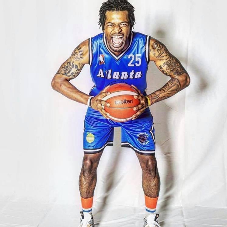 Man in blue basketball uniform holding a basketball, smiling, with tattoos on arms, against a white background.