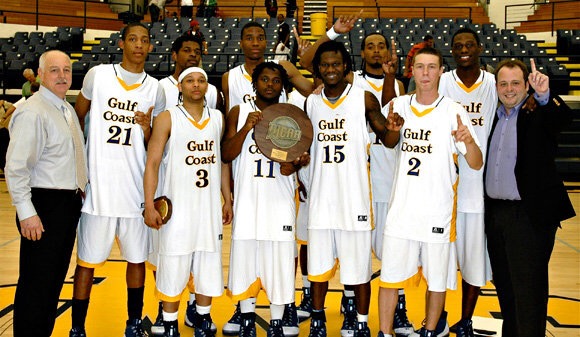 Group of basketball players and coaches posing with a trophy on a court, some holding medals, in a sports arena.