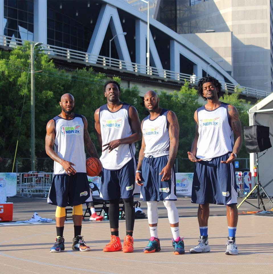 Four basketball players in uniform standing outdoors on a court with modern buildings in the background.