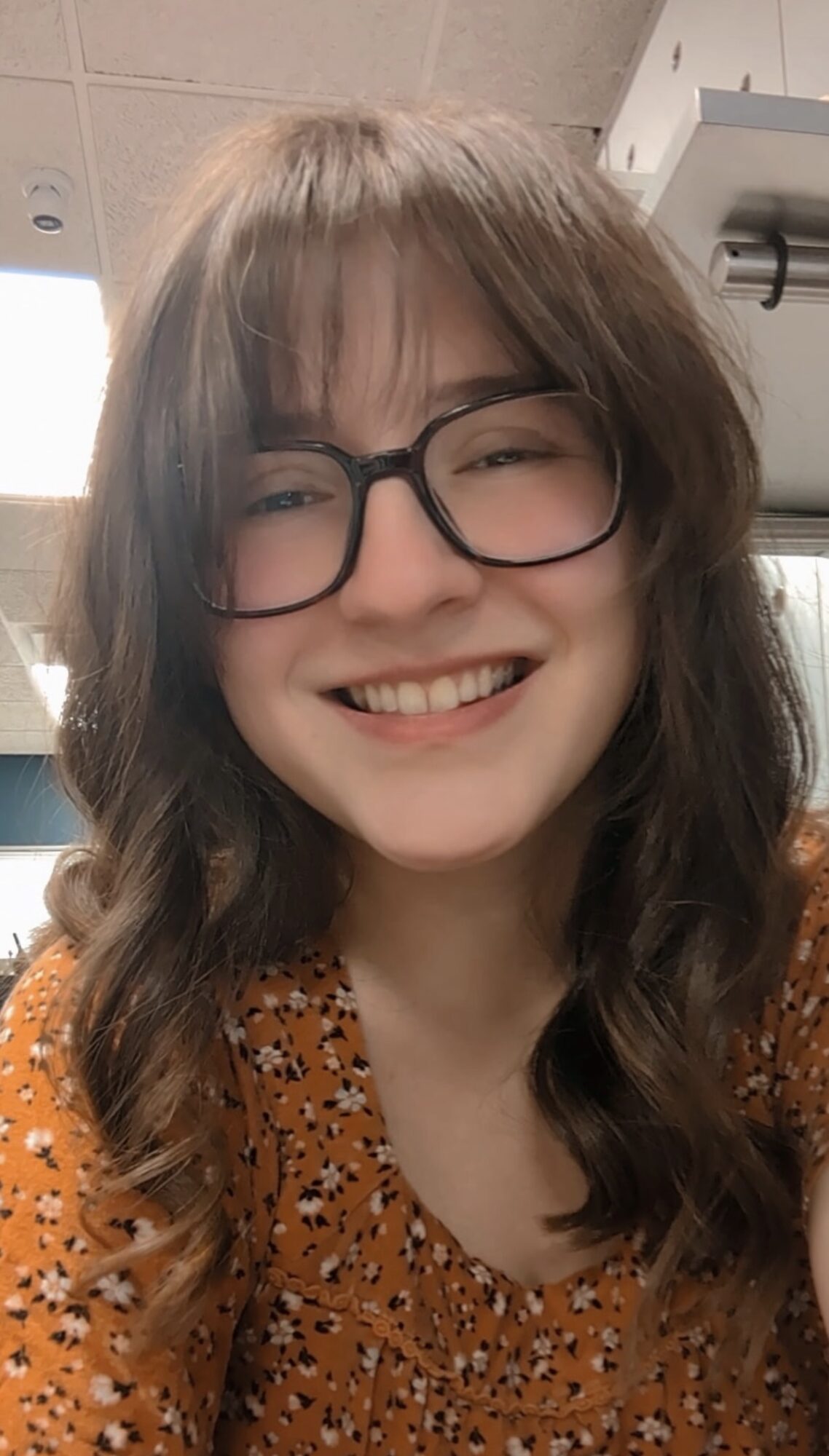 Young woman with glasses and wavy hair smiling, wearing an orange patterned top, indoors.