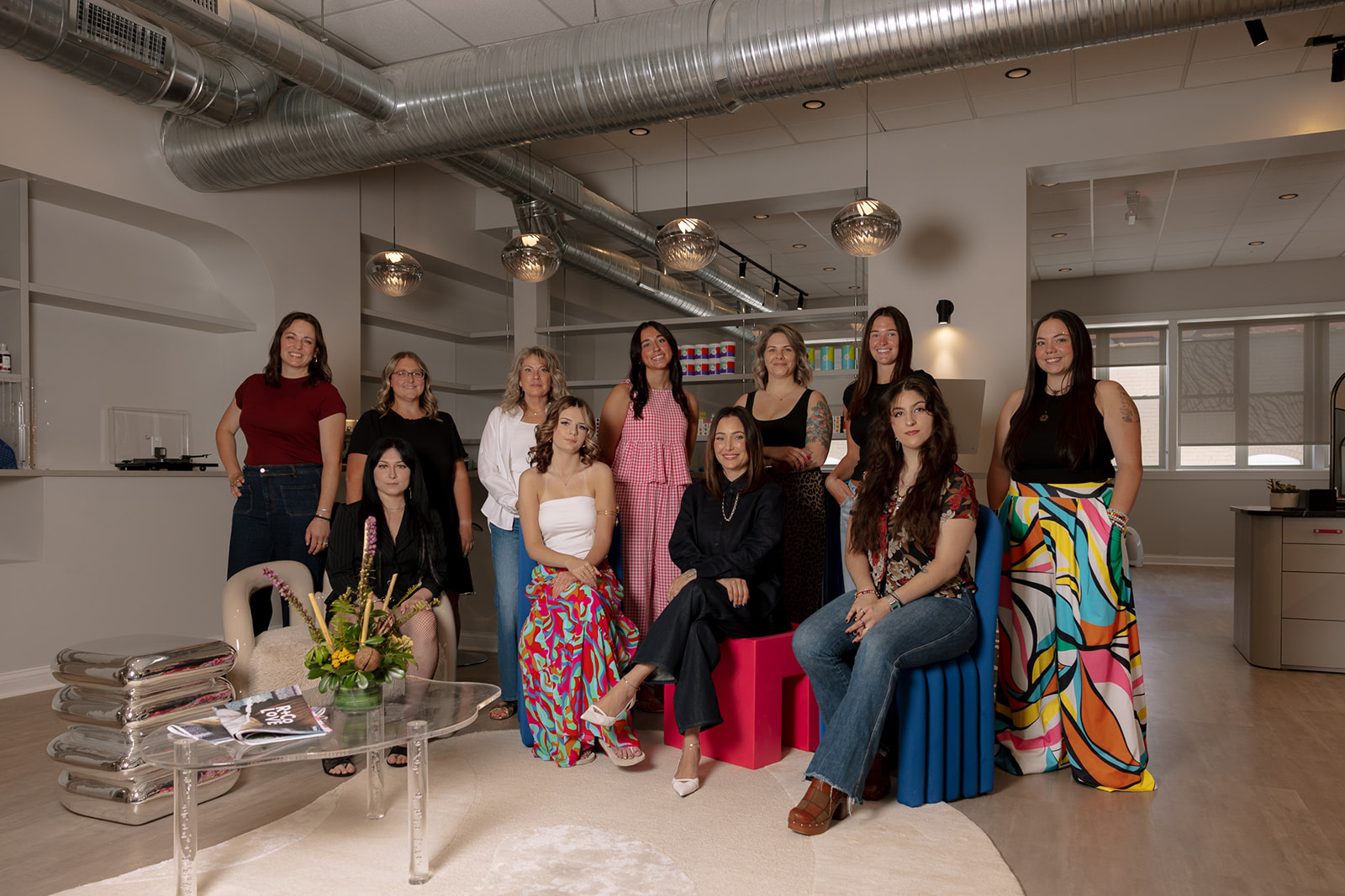 Group of ten women in a modern, well-lit room with industrial ceiling, some sitting and some standing, dressed in colorful and casual clothing.