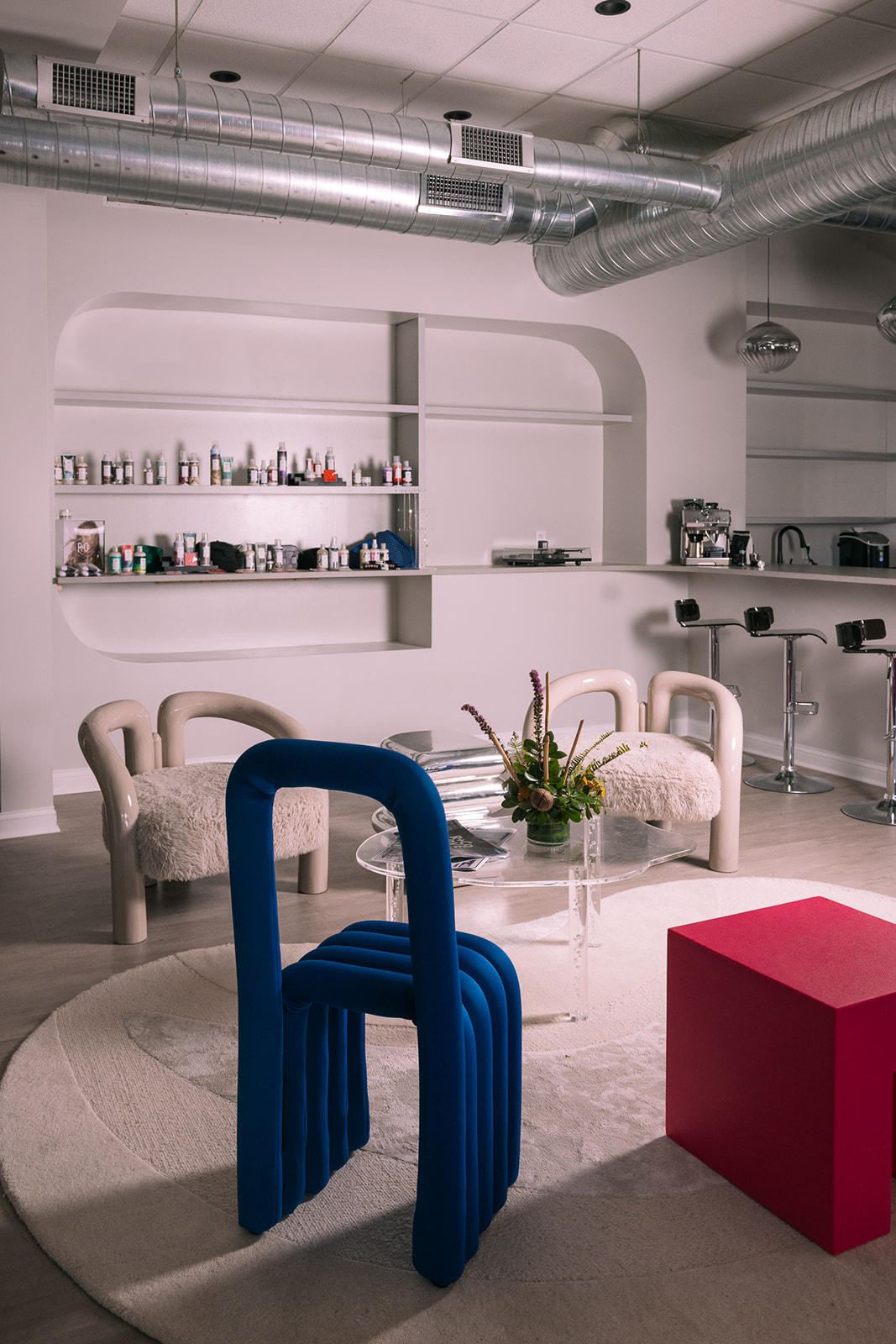 Modern interior with white walls, shelves, seating, a round rug, and colorful furniture including blue chairs and a red block.