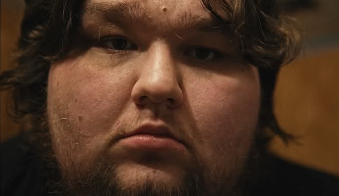 Close-up of a person's face with a serious expression, dark hair, and facial hair.
