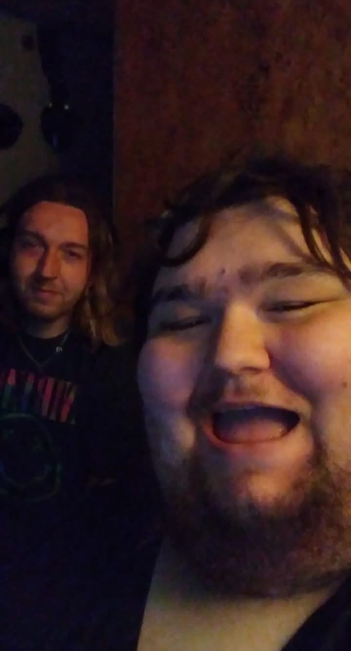 Two people smiling, one in the foreground and one in the background, in a dimly lit setting.
