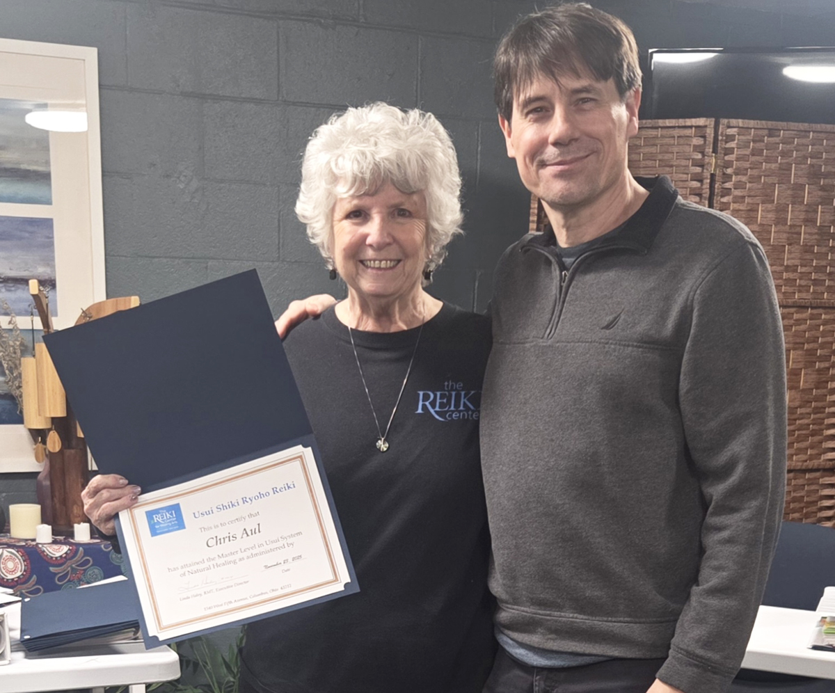 Two people, a woman with white curly hair and a man with dark hair, stand indoors. The woman holds a certificate and smiles.