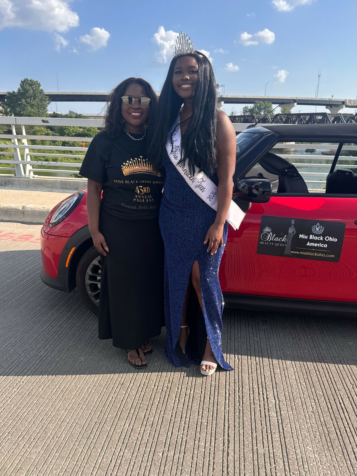 Two women standing outdoors near a red car, smiling, with a cityscape and blue sky in the background.