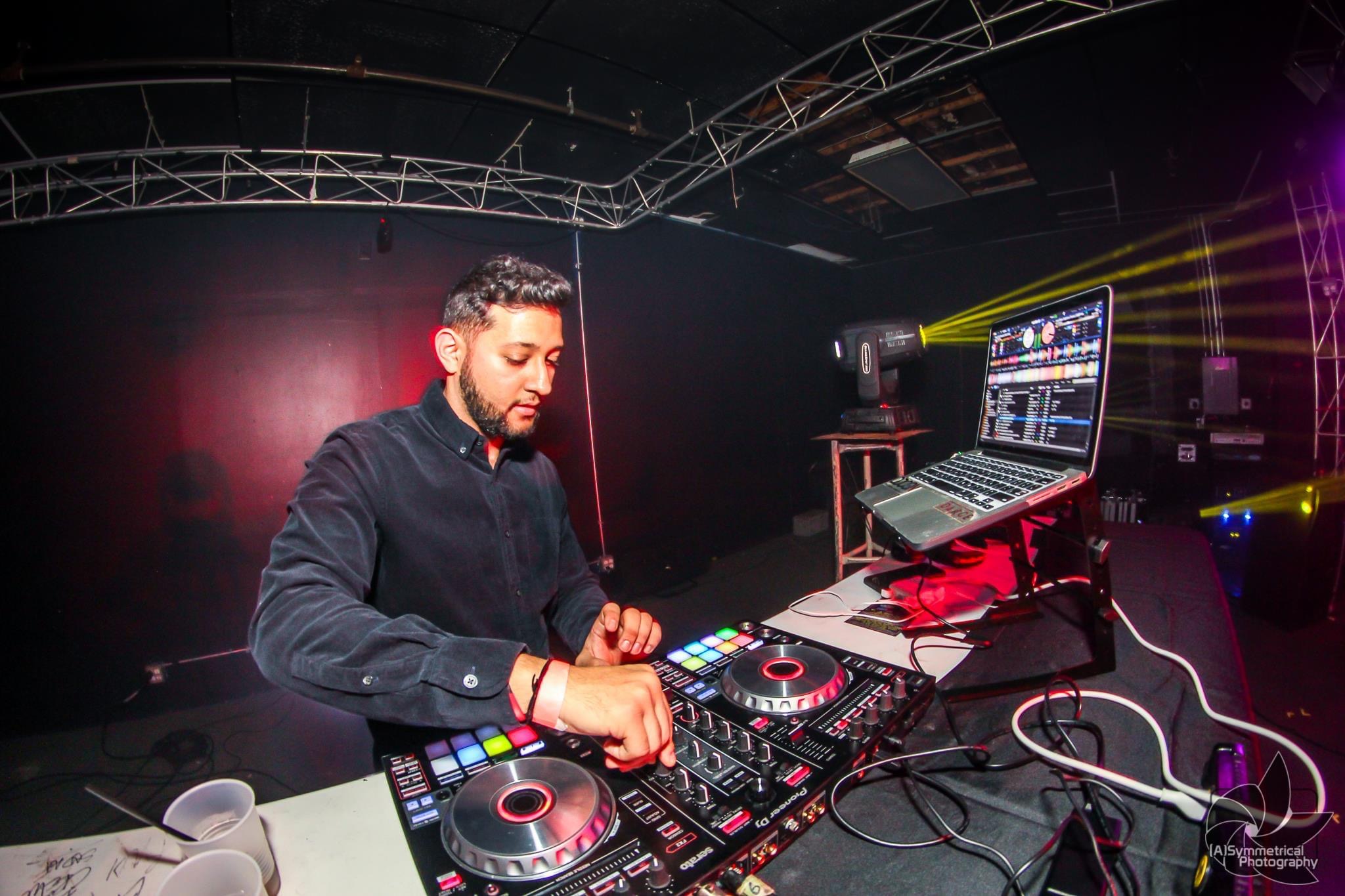 DJ with equipment in a dark club, colorful lights, and a large screen displaying DJ software, focused on mixing music.
