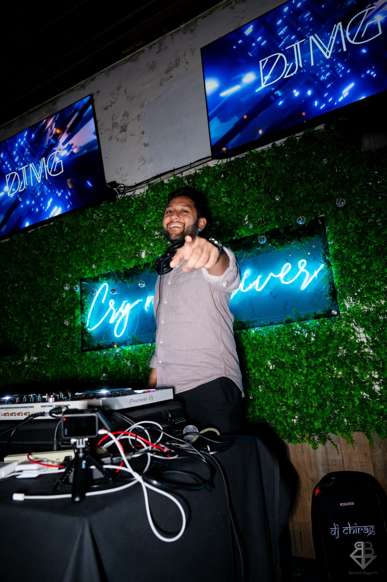 DJ with beard pointing at camera, standing in front of green wall with neon signs, DJ equipment on table.
