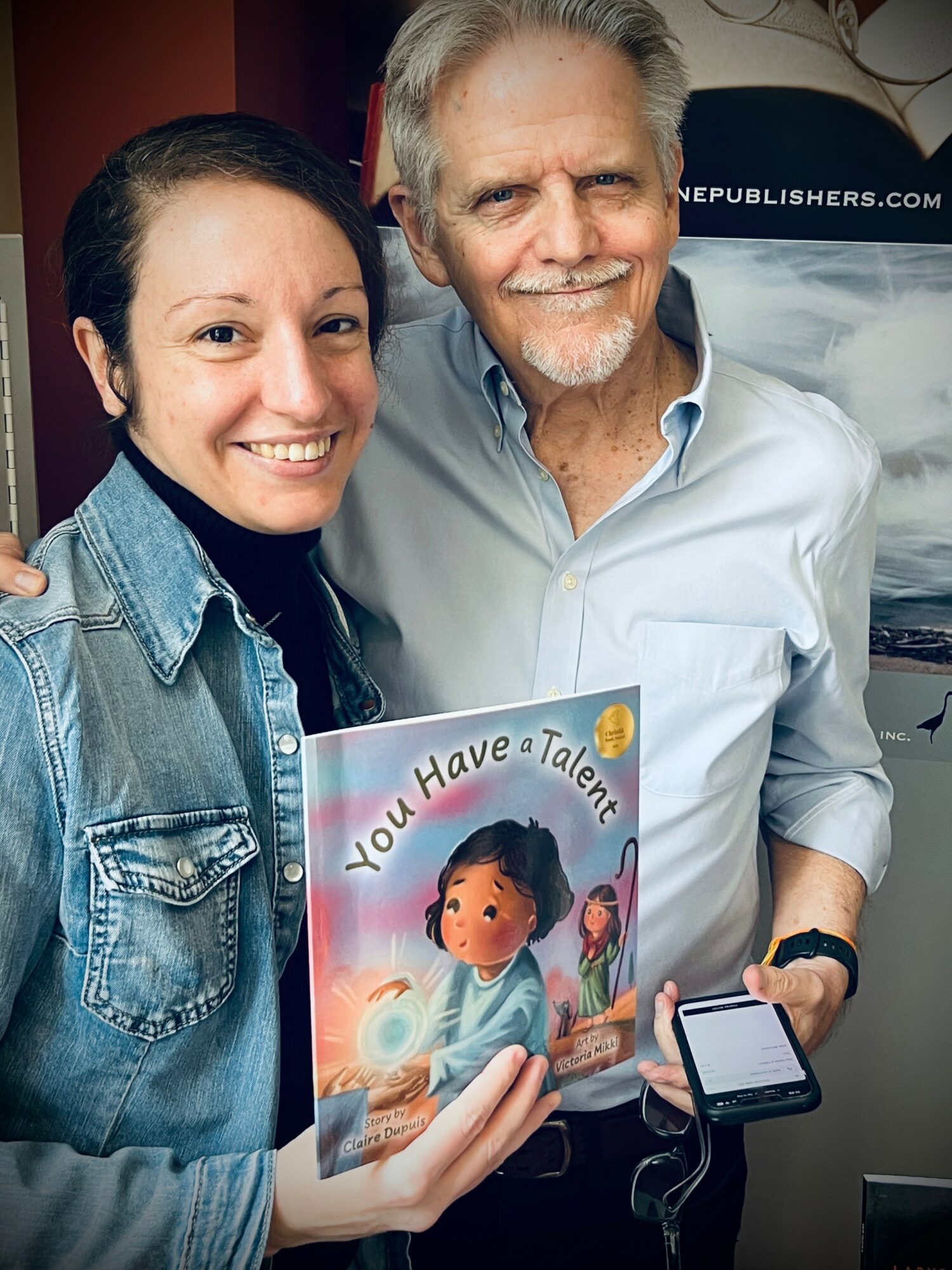 Two smiling people holding a children's book titled 'You Have a Talent,' with a woman holding a phone, in an indoor setting.