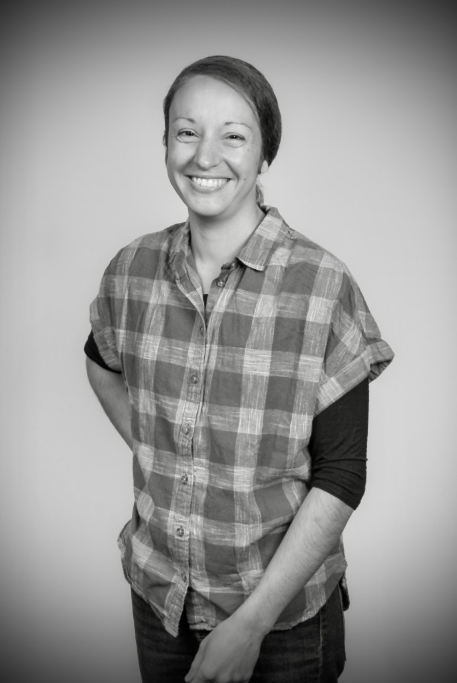 Smiling woman with short hair wearing a checkered shirt and black sleeves, standing against a plain background.