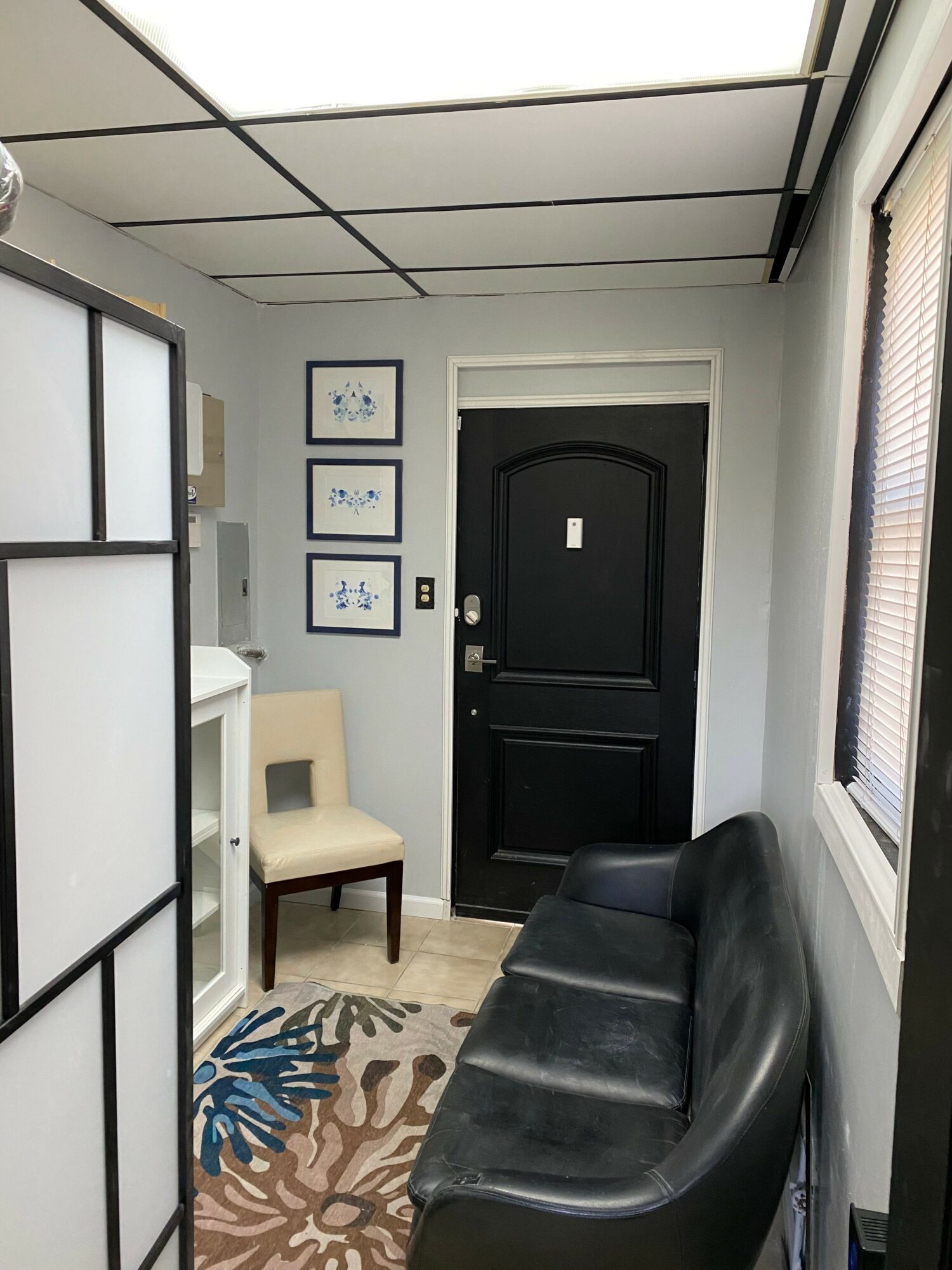View of a room with a black door, framed pictures, a beige chair, a black sofa, and a patterned rug.